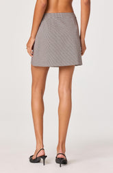 A woman is wearing a high-waisted, A-line mini skirt with a small checkered pattern in neutral colors. She is also wearing black high-heeled shoes with ankle straps. The image shows her from the back, with her bare upper body visible.